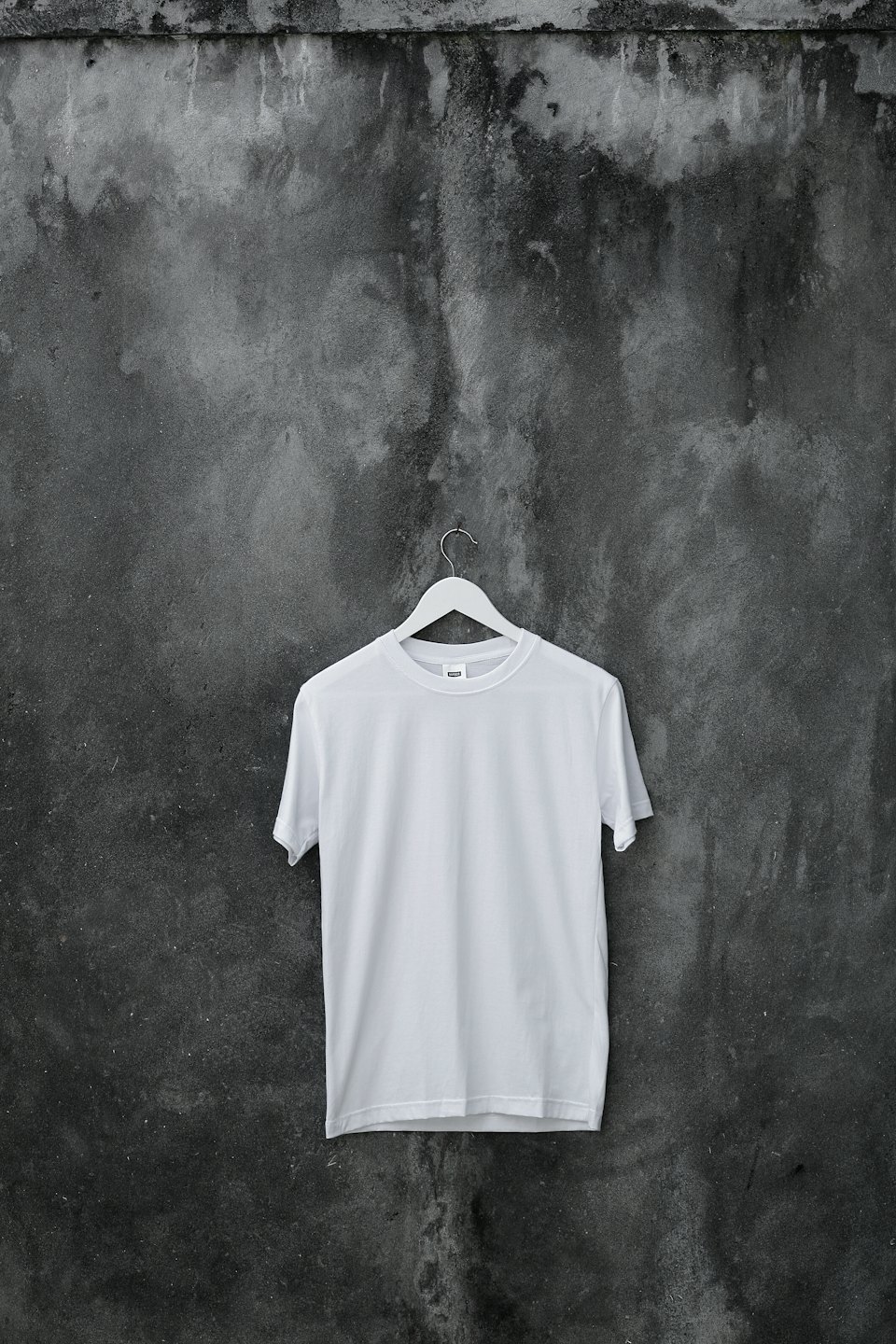 A white t-shirt hangs on a hanger against a concrete wall.